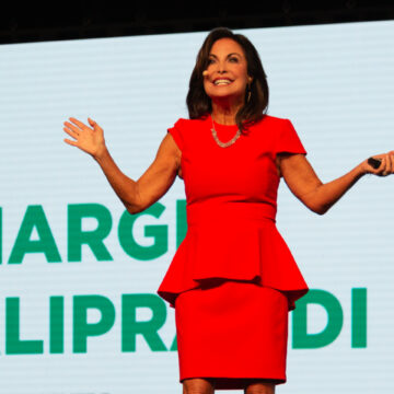 Event-Photography Margie Aliprandi presenting on stage at the Modere 20/20 Vision conference, wearing a vibrant red dress, with her name displayed on a large screen behind her, captured by Coldea Productions