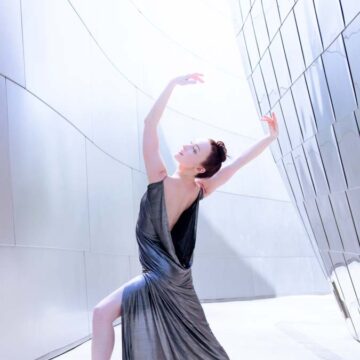 A dancer in an elegant silver dress striking a graceful ballet pose against the modern, metallic architecture of the Walt Disney Concert Hall in Los Angeles.