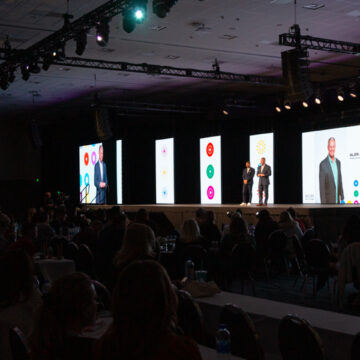 Convention-Photography Audience at a live event viewing a presentation on stage with large screens displaying Alan Jones' profile and colorful visuals.