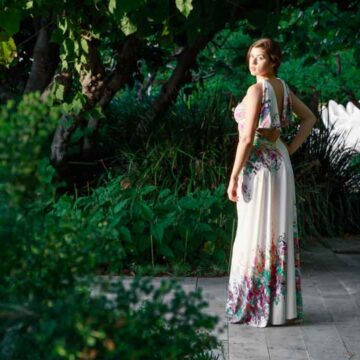A woman in an elegant, floral dress posing outdoors among lush greenery, demonstrating fashion and portrait photography.