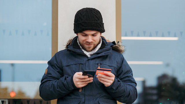 A smiling man in a jacket using a smartphone and holding a credit card