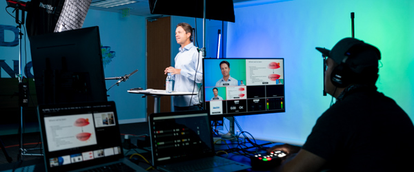 A speaker delivering a presentation in a studio while a video technician manages the live broadcast using multiple monitors.