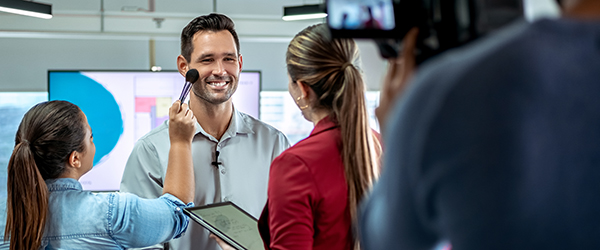 Behind-the-scenes of a video shoot featuring a smiling man being prepped with makeup and assisted by a production team.