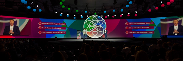 A large stage with colorful geometric shapes and designs, featuring a presenter speaking to a large audience.