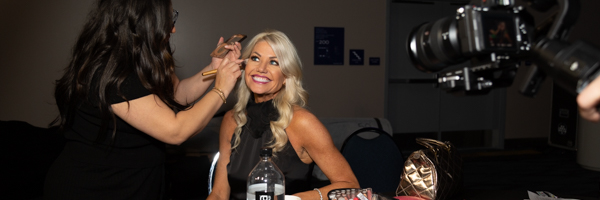A makeup artist preparing a smiling woman for an event, with a professional camera capturing the moment.