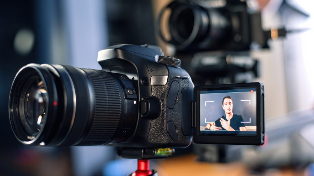 Close-up of a DSLR camera recording a man giving a presentation on its flip screen. The man is seated in front of the camera, speaking with hand gestures, while another camera is positioned in the background.