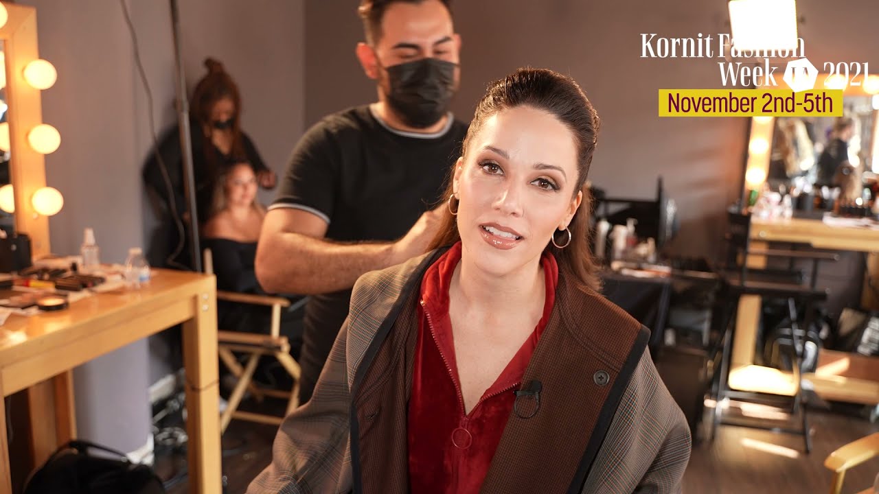 Backstage daily presentation at Kornit Fashion Week LA 2021 featuring Steward, who is in the makeup and styling area. Steward, wearing a red top with a stylish jacket, is seated in front of a well-lit mirror while a makeup artist prepares her for the day's events. The logo and dates for Kornit Fashion Week, "November 2nd-5th," are displayed in the top right.