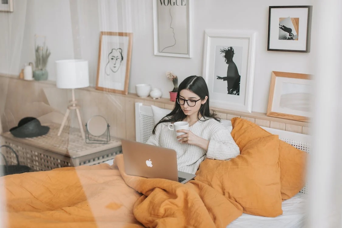 A woman drinking tea while lying in bed with a laptop.