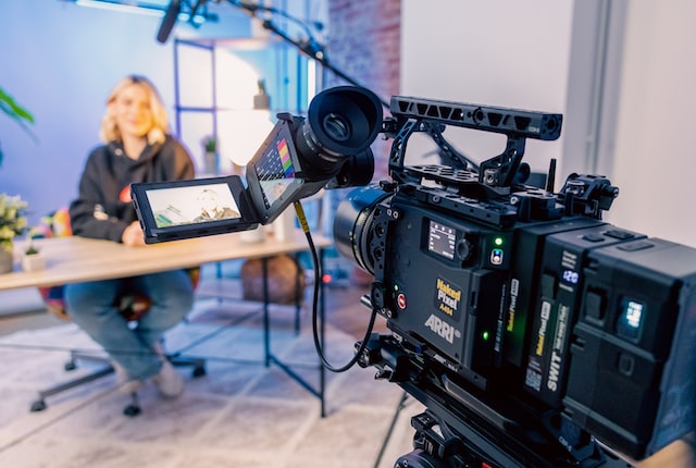 Close-up of a professional ARRI camera setup in a studio recording a person seated at a table, with video monitors displaying the subject in the frame.
