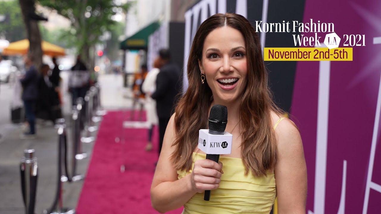 A cheerful host introduces Kornit Fashion Week LA 2021 on the first day of the event, standing on the pink carpet with a microphone in hand. The event’s branding and dates, November 2nd-5th, are visible in the background.