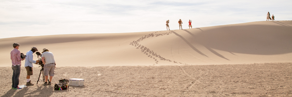 Production crew filming on a vast desert landscape with footprints leading over a sand dune, used as the page header for the Commercials Video Production in Palm Springs, California geolocation service page