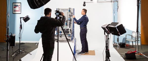 Rob Gledhill of Blue-White Industries demonstrates the functionality of a Plastic Chemical Feed Skid while Alexis Mendez from Coldea Productions films the product presentation video.