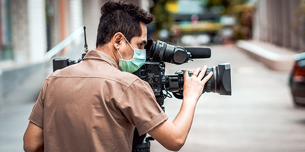 A videographer wearing a face mask operating a professional video camera on a production set outdoors.