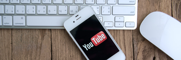 A smartphone displaying the YouTube logo placed on a wooden desk alongside a keyboard and mouse, representing Coldea Productions' web video production services.