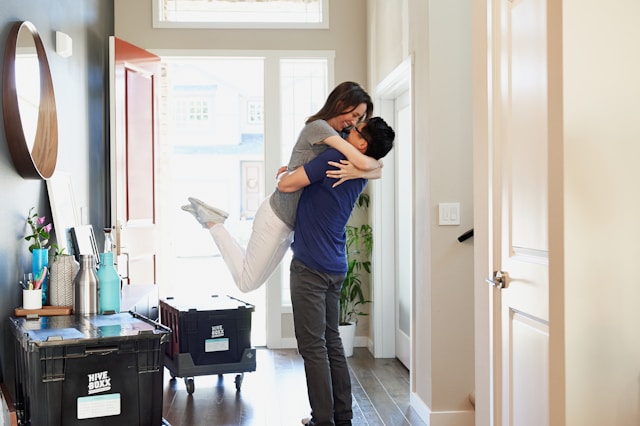 Woman in gray long-sleeve shirt and gray pants standing beside a white wooden door, hugging her spouse.