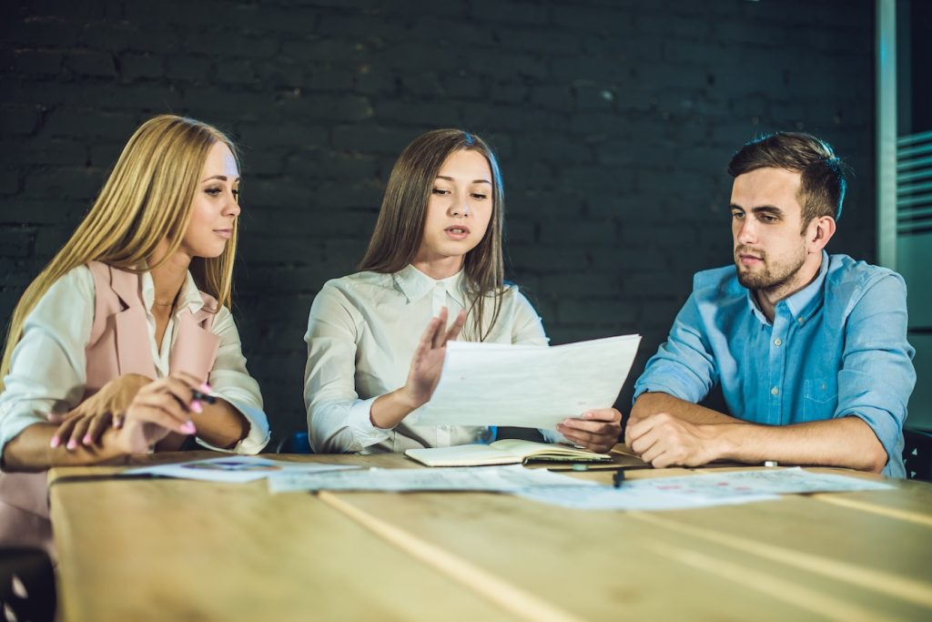 A group of three people sitting at a table, with one person holding a document and discussing it, symbolizing collaboration and script review in video production with the guidance of a writing coach.